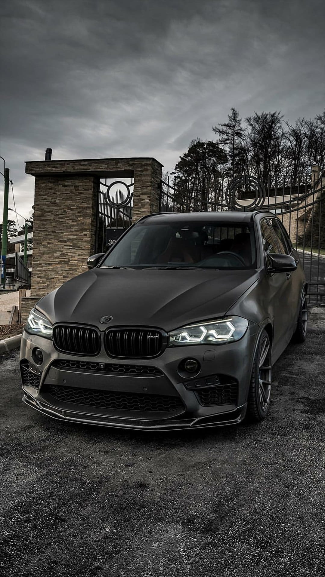 BMW X5 SUV in matte black finish parked on asphalt with LED lights glowing against a cloudy sky and stone pillars.