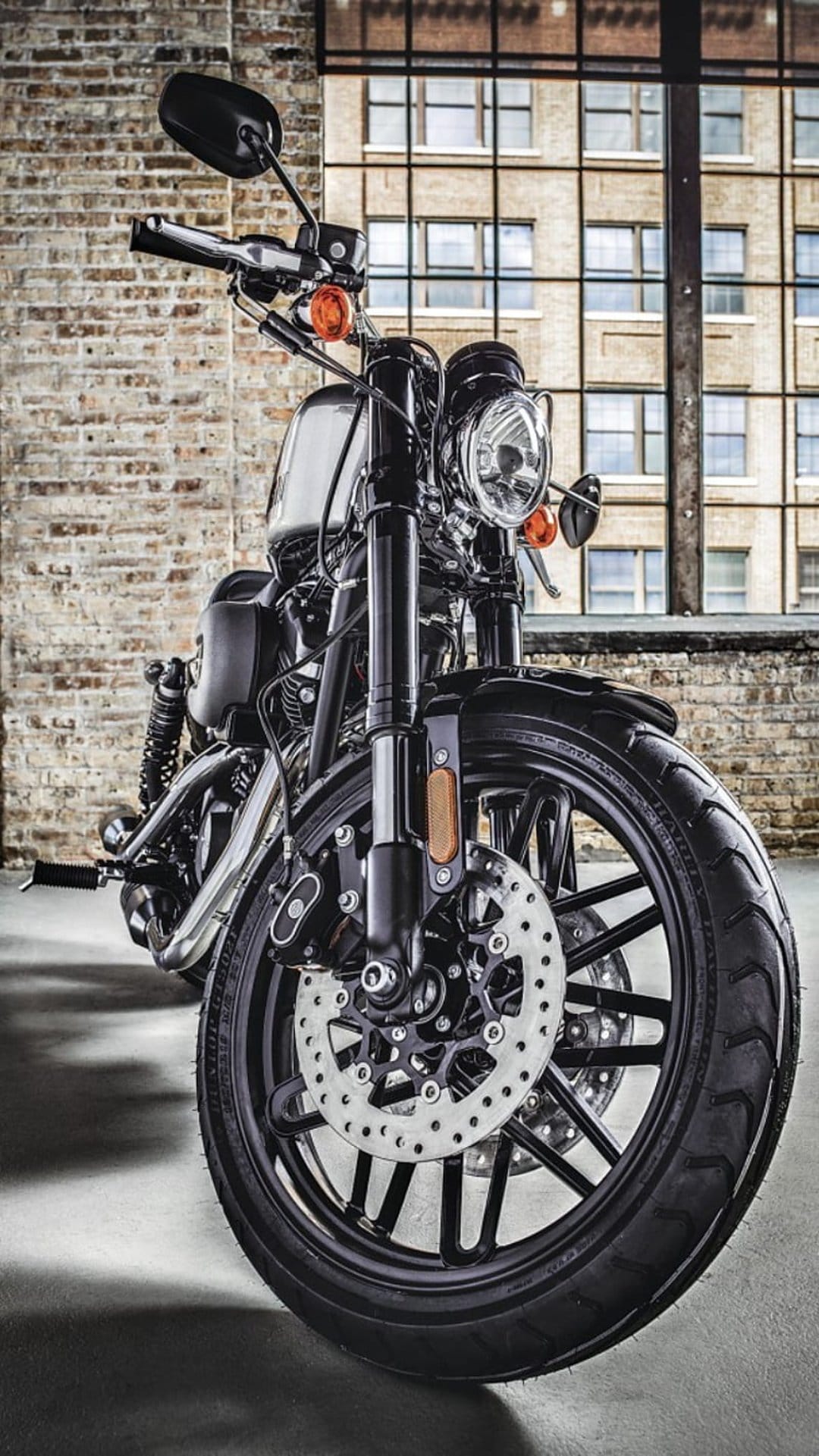 Harley Davidson front wheel and headlight against a brick wall with industrial windows in an urban setting.
