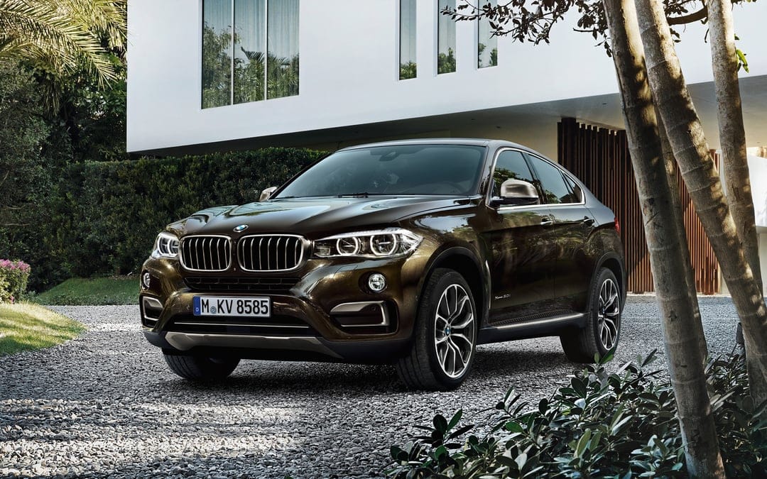 Brown BMW X6 parked on a gravel driveway in front of a modern white house with large glass windows and green trees.