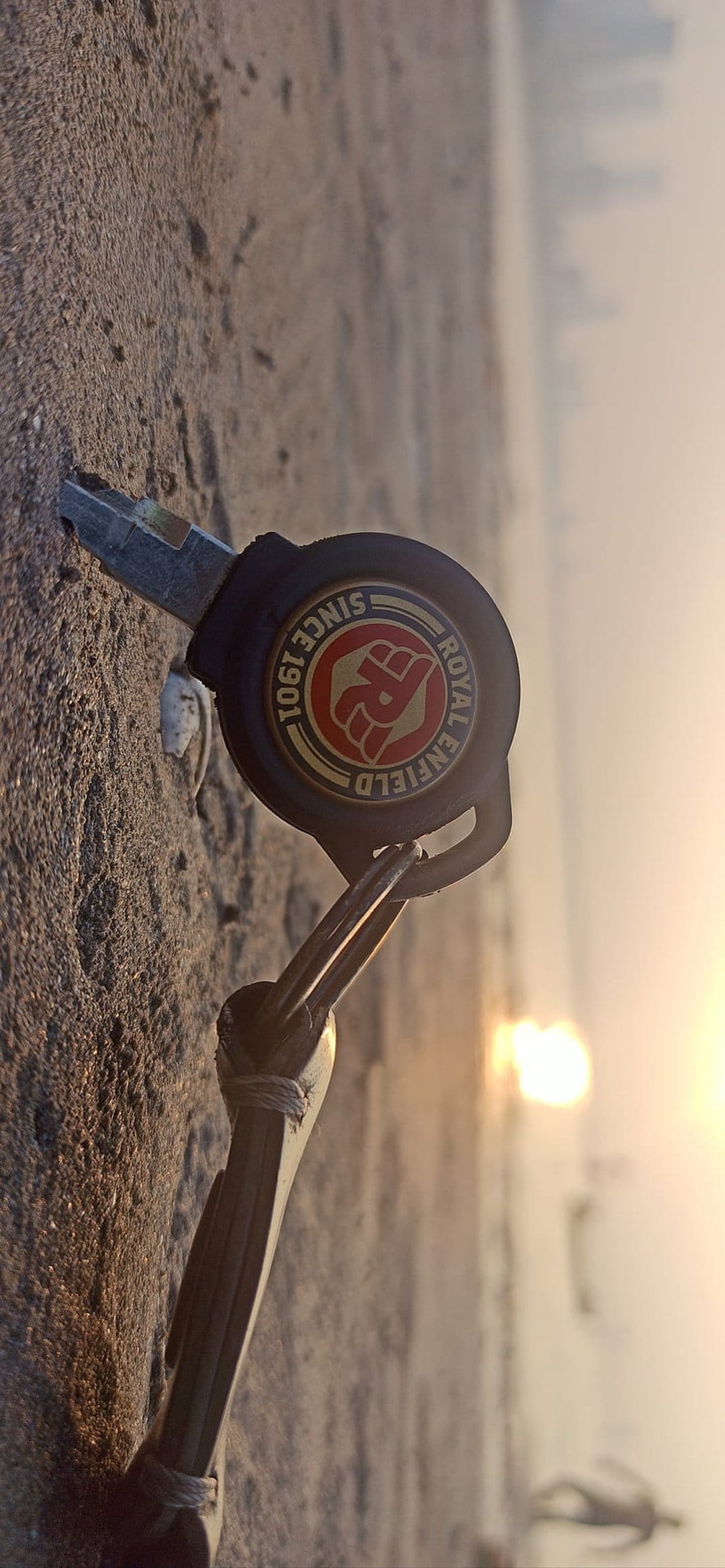 Royal Enfield key on sandy beach with a golden sunrise over the ocean and a distant city skyline background.