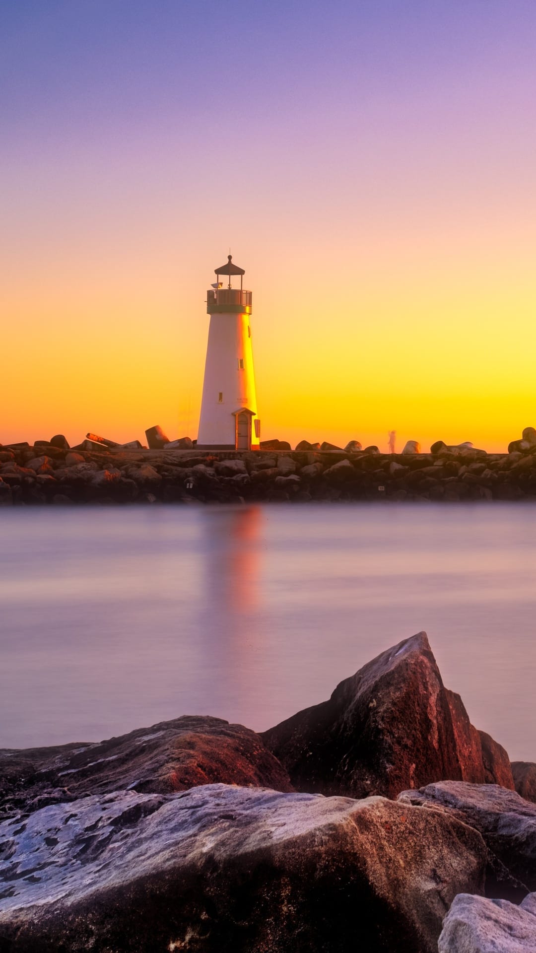 White lighthouse on dark rocks under a purple and orange sunset sky reflecting in calm coastal water.