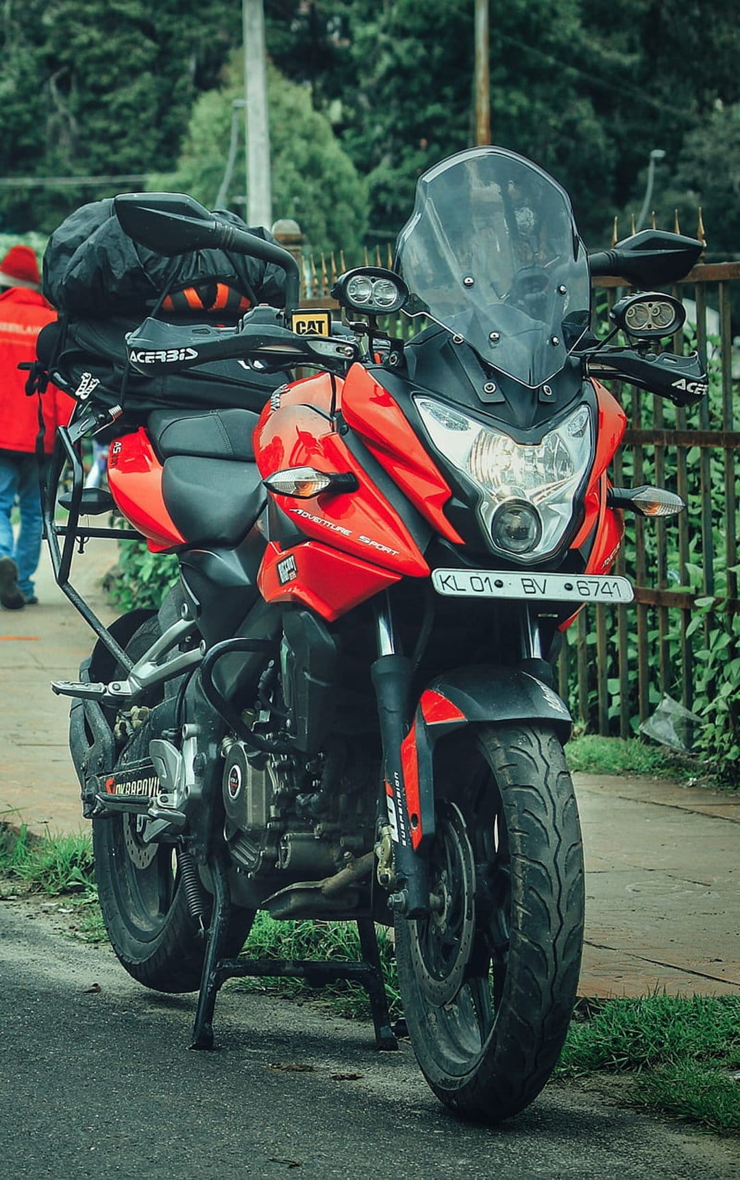 Red adventure motorcycle with a black luggage bag parked on a rural roadside next to lush green foliage.