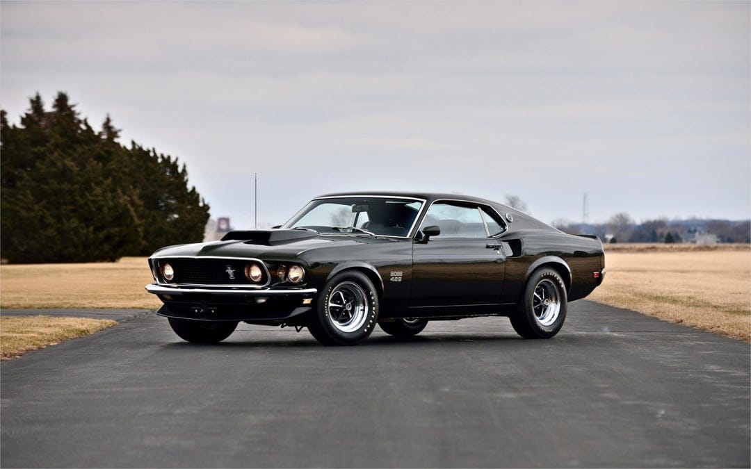 Dark green 1969 Ford Mustang Boss 429 muscle car on a paved road against a muted sky and vast dry fields.