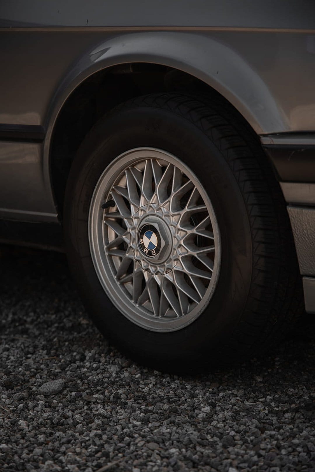 BMW star-spoke alloy wheel with a central logo sits on a gravel surface against a metallic gray car body.