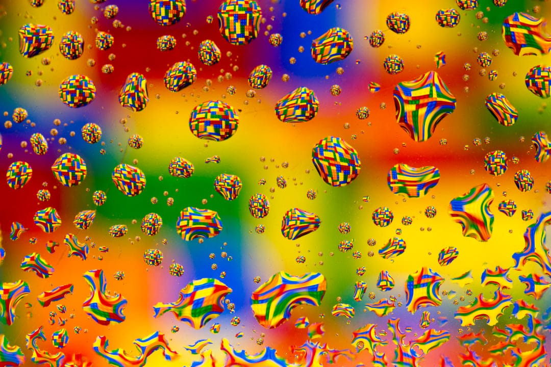 Translucent water droplets resting on a multi-colored surface reflect a vivid rainbow gradient in sharp focus.