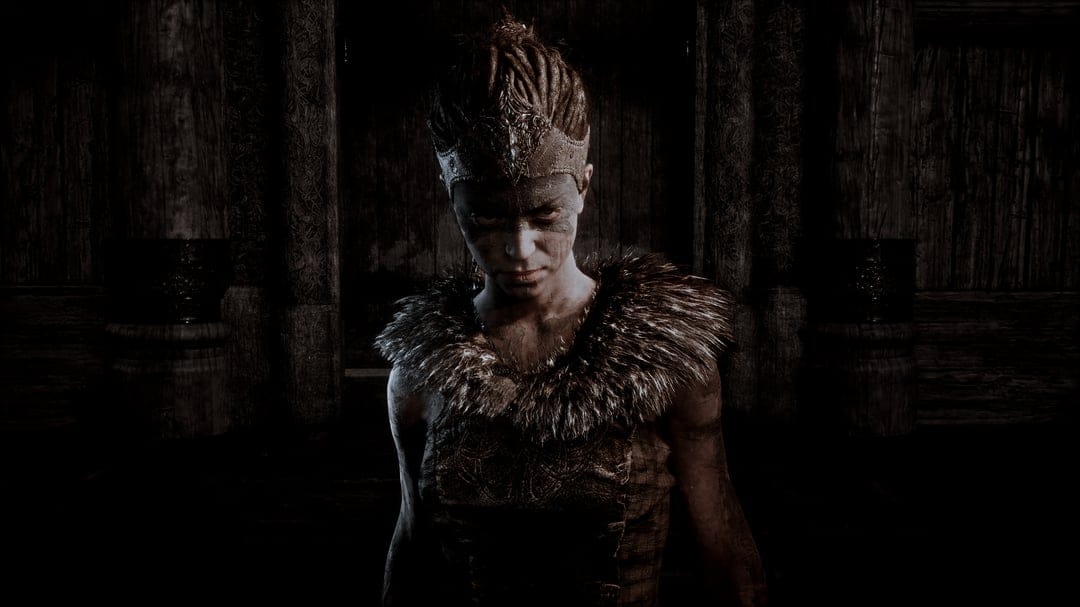 Senua with face paint and fur collar stares intensely in a dark wooden Norse interior with moody lighting.