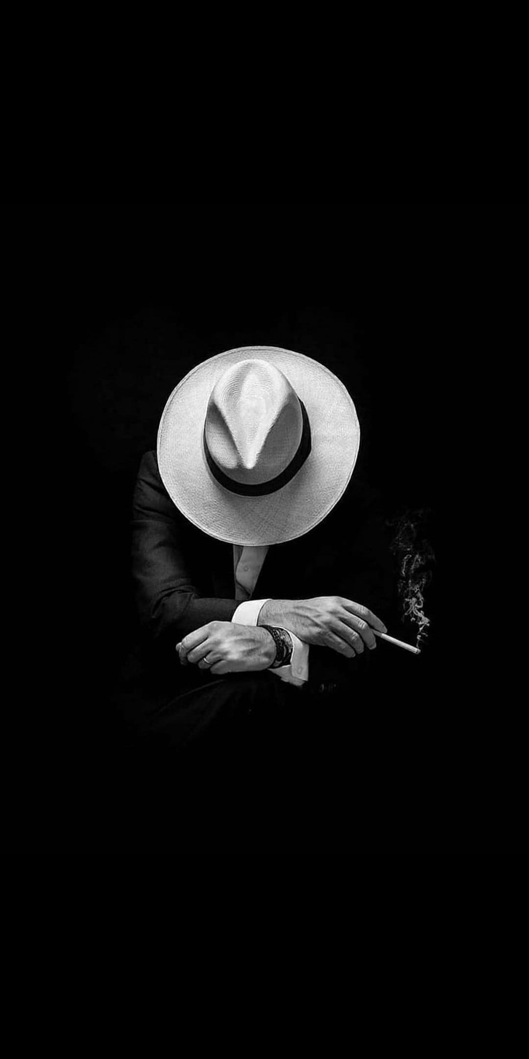 Man in a suit and fedora holds a glowing cigarette with smoke rising against a dark, high-contrast background.