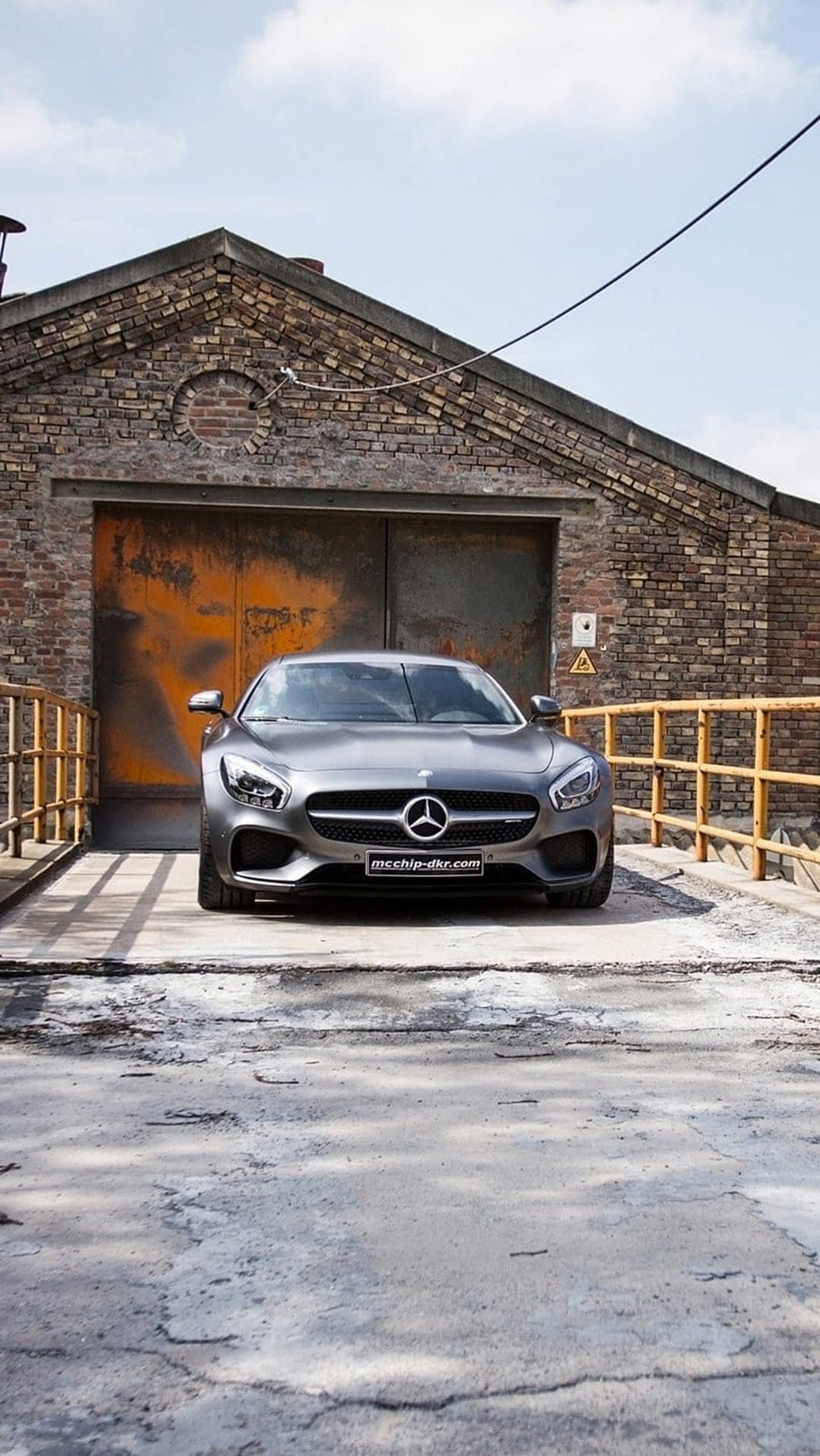 Gray Mercedes-AMG GT coupe parked on a paved driveway in front of a weathered brick warehouse with rusted doors.