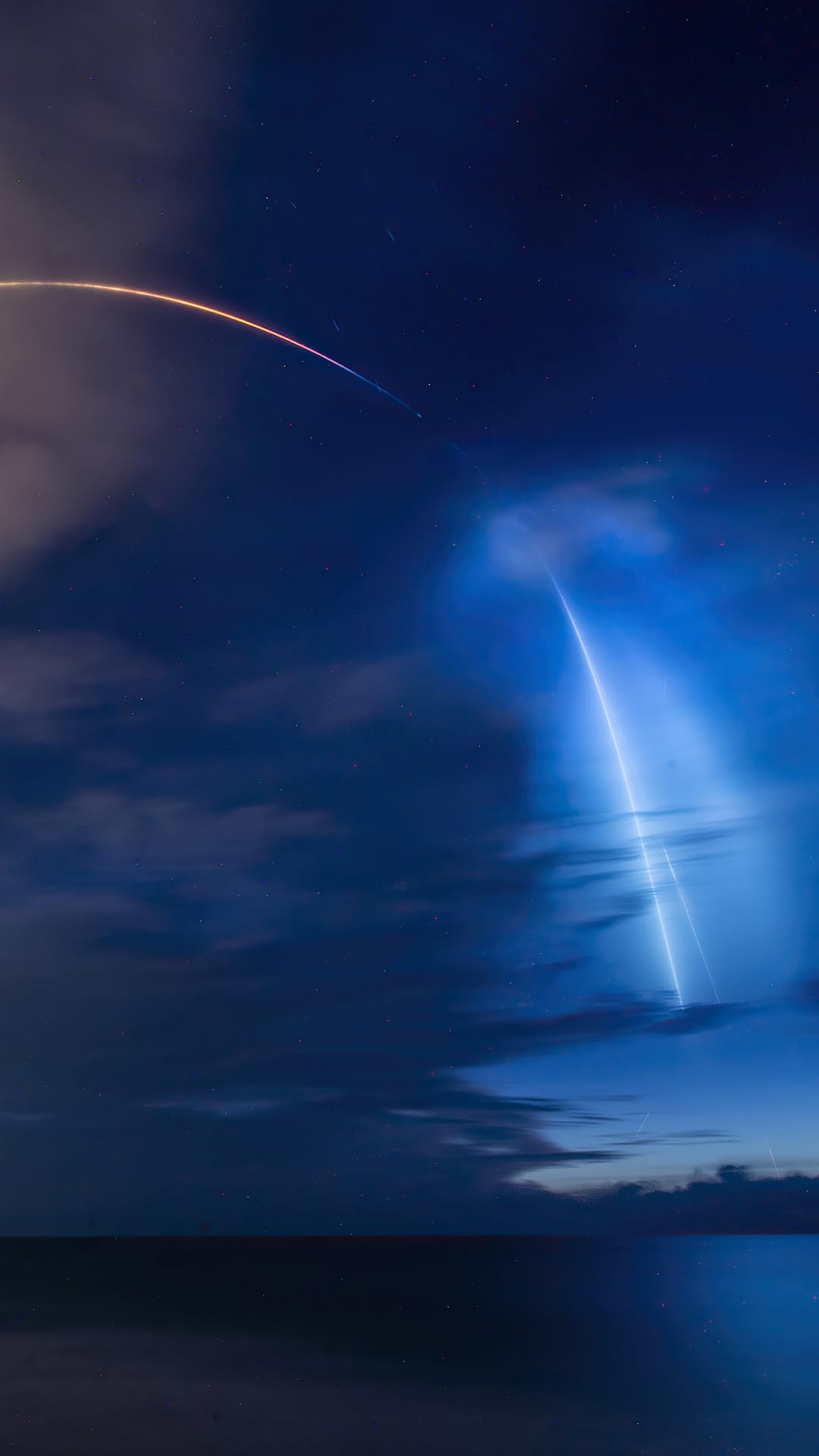 Rocket launch plume glowing blue above a dark ocean horizon with a bright white light trail in the night sky.