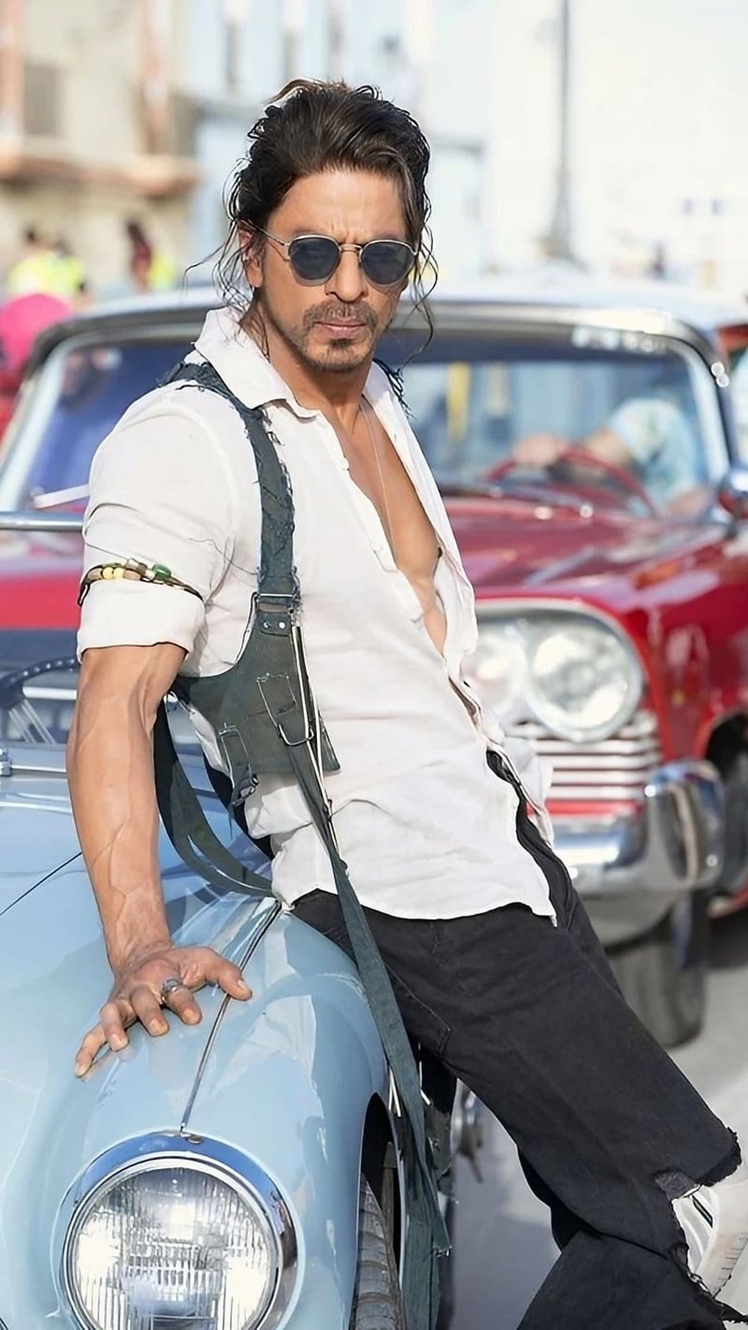 Shah Rukh Khan leans on a light blue vintage car wearing a white shirt, harness, and sunglasses in a retro setting.