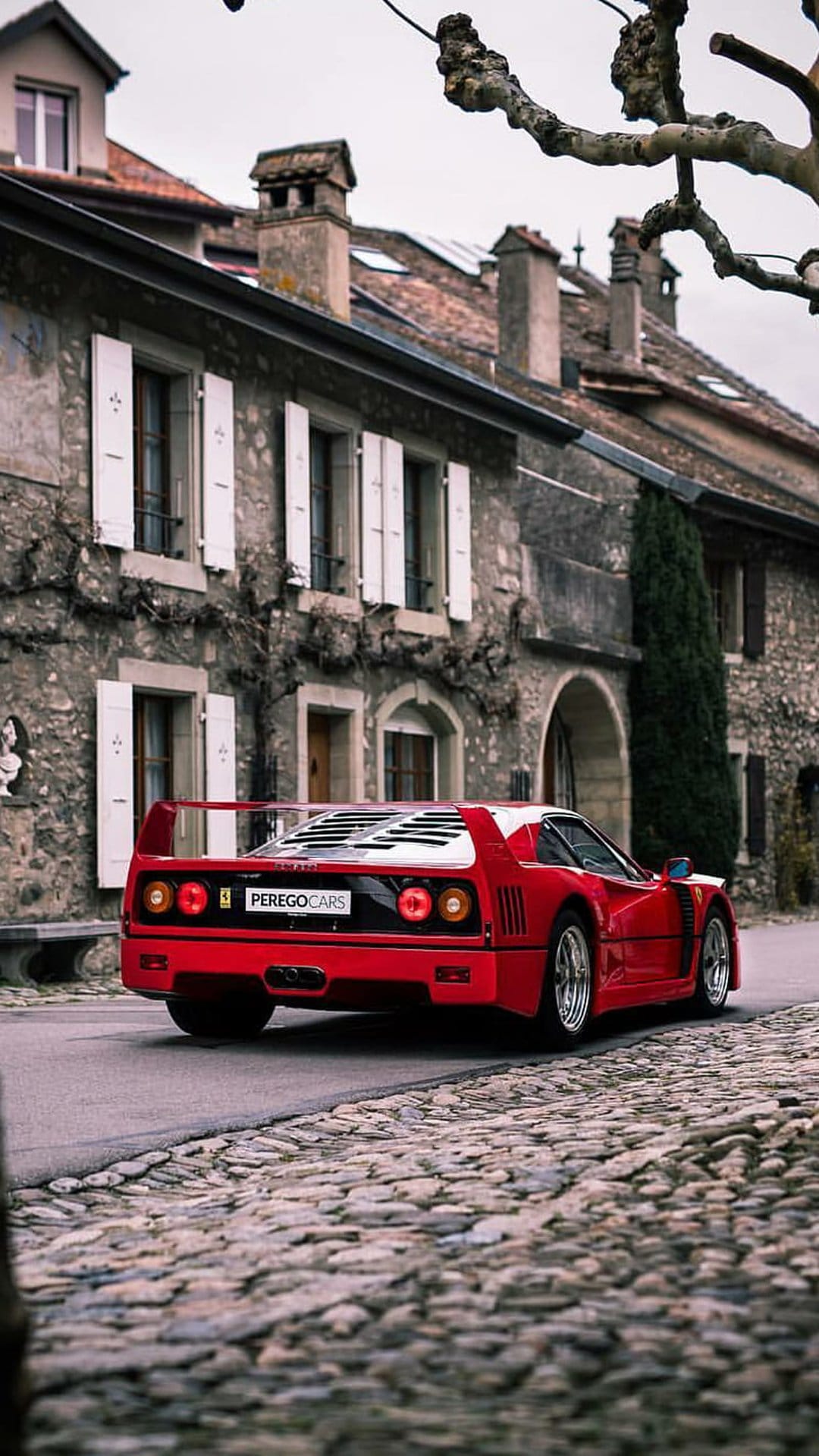 Red Ferrari F40 supercar parked on a rustic cobblestone street lined with historic stone buildings and shutters.