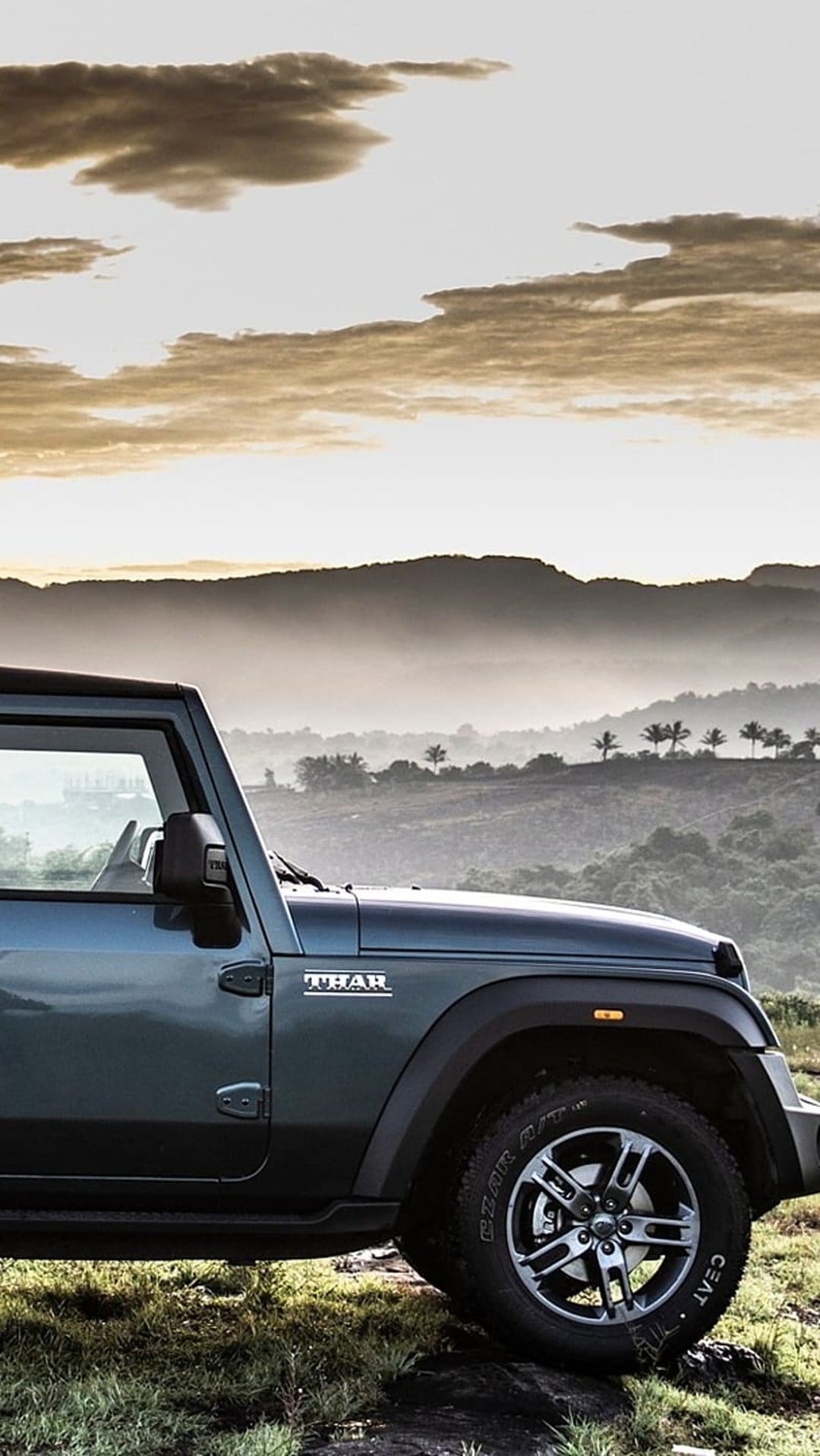 Dark blue Mahindra Thar SUV parked on a grassy hill with hazy mountains and a sunset sky in the background.