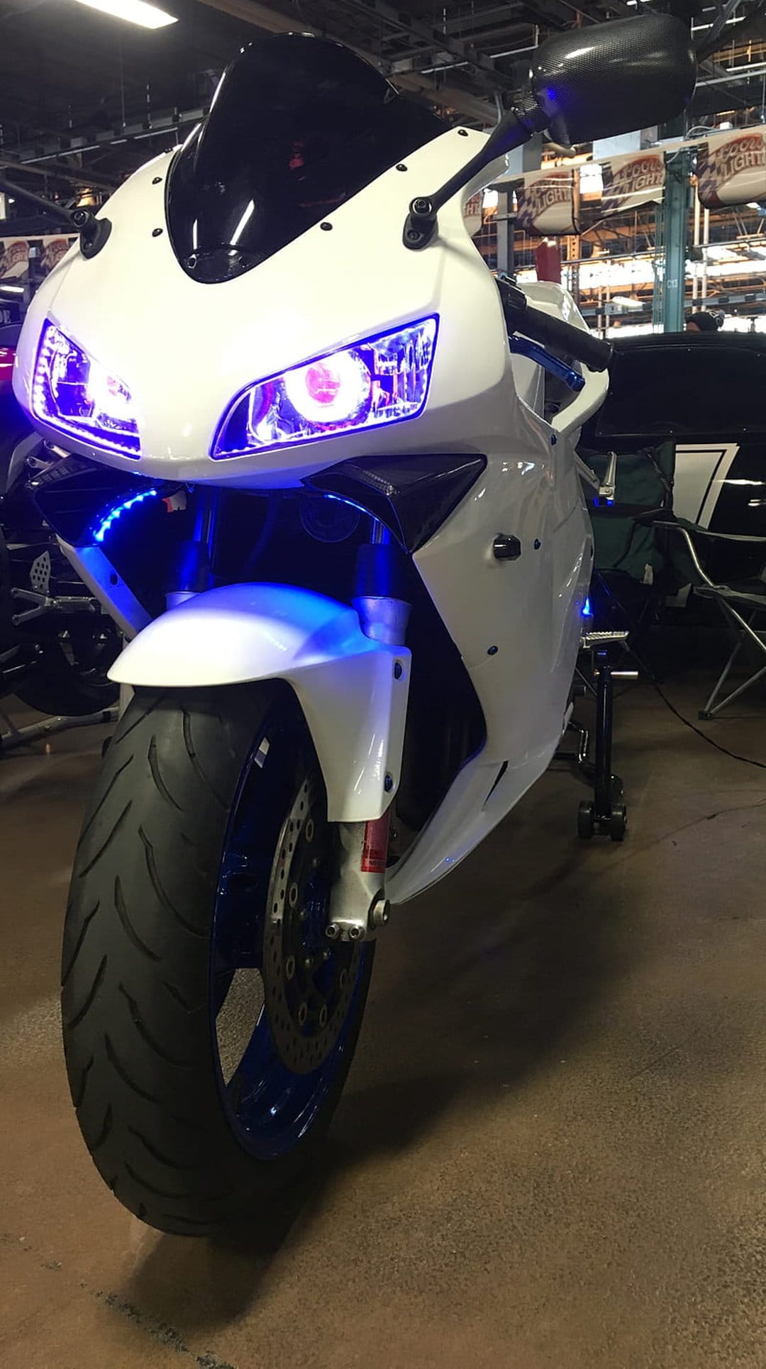 White sportbike with vibrant blue LED underglow and headlights on a dark floor in a vertical composition.