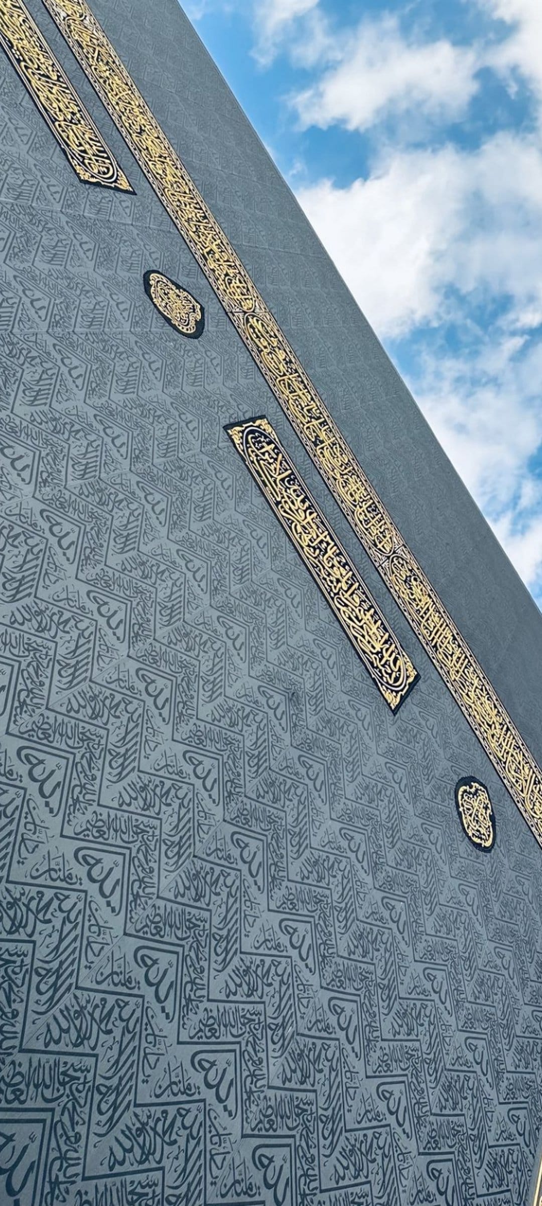 Gold Arabic calligraphy embroidered on the black silk Kiswah fabric of the Kaaba against a blue sky background.