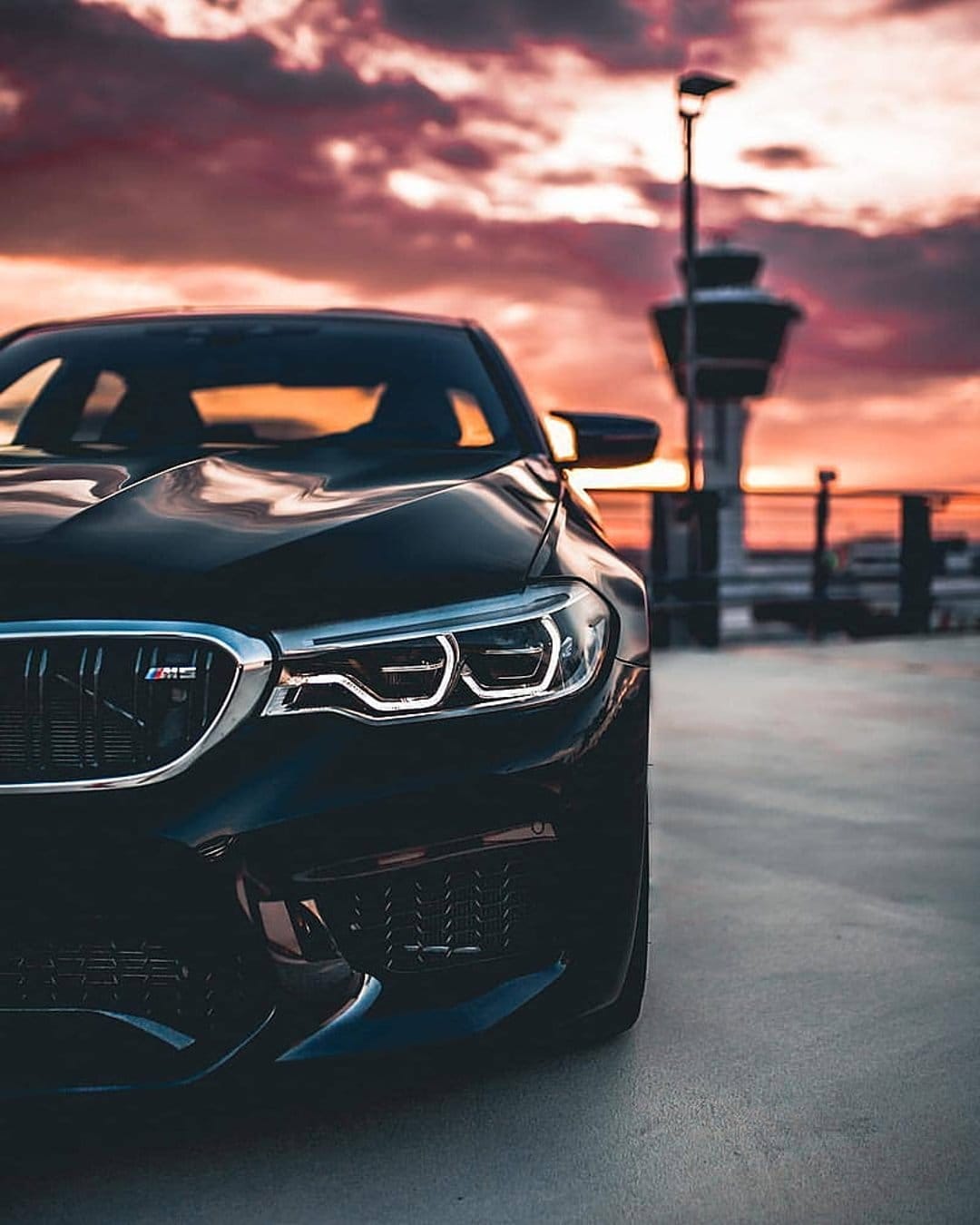 Black BMW M5 sport sedan with lit headlights parked at twilight near an airport control tower silhouetted by clouds.