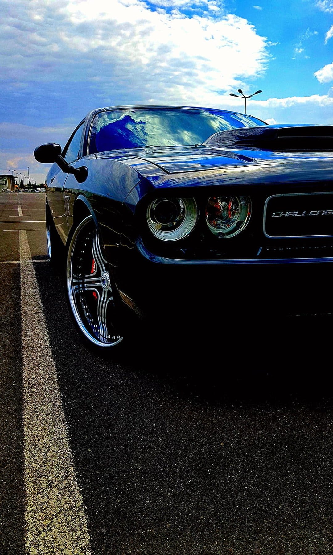 Dark blue Dodge Challenger R/T parked on asphalt with chrome wheels reflecting a bright cloudy sky at low angle.