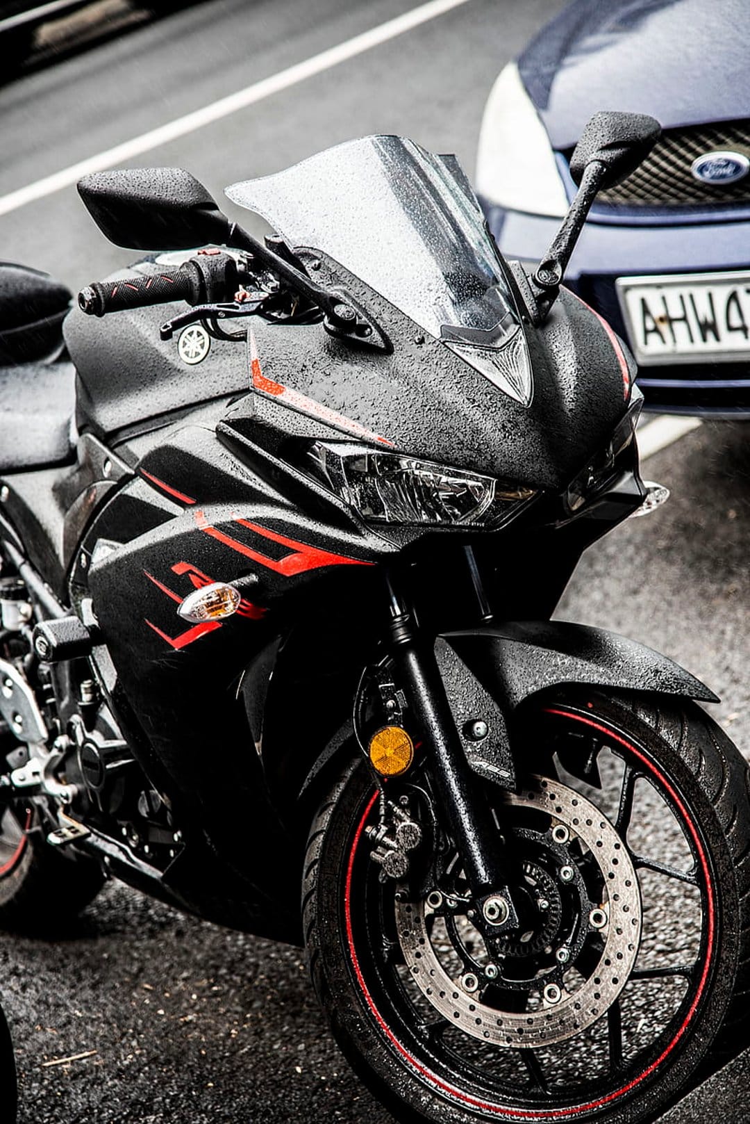 Black sport motorcycle with red rims and wet fairing parked on damp asphalt during a rainy day near a blue car.