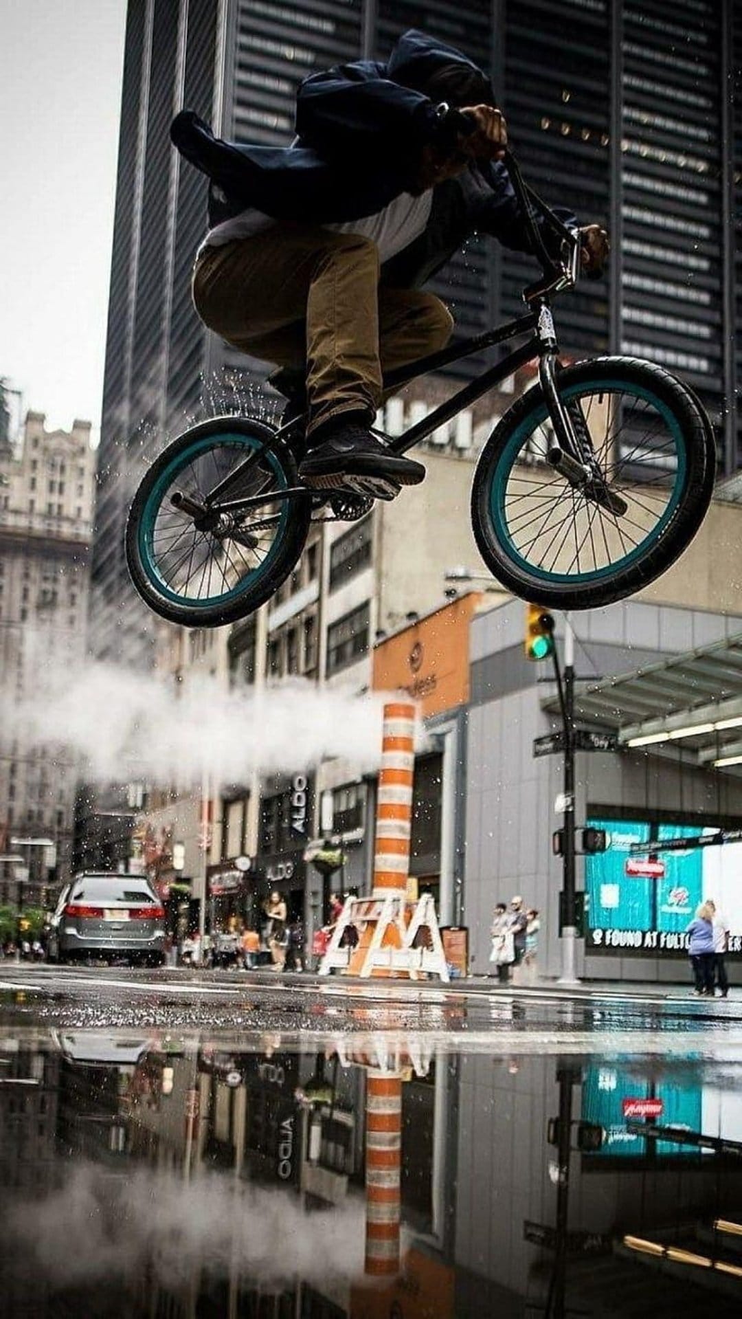BMX rider jumps over white steam rising from a wet city street with building reflections in rain puddles.