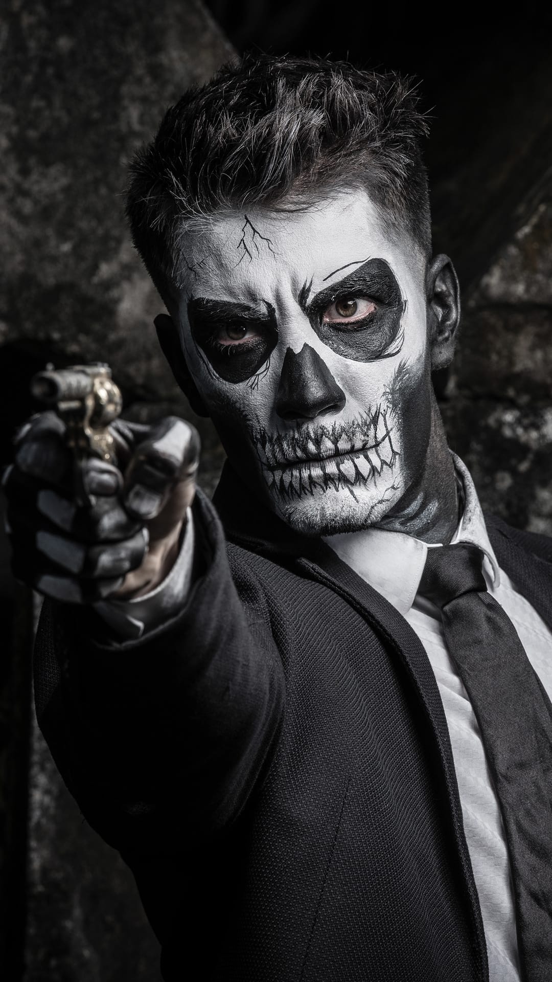 Man in a black suit with skull face paint aims a handgun forward against a dark, moody background.