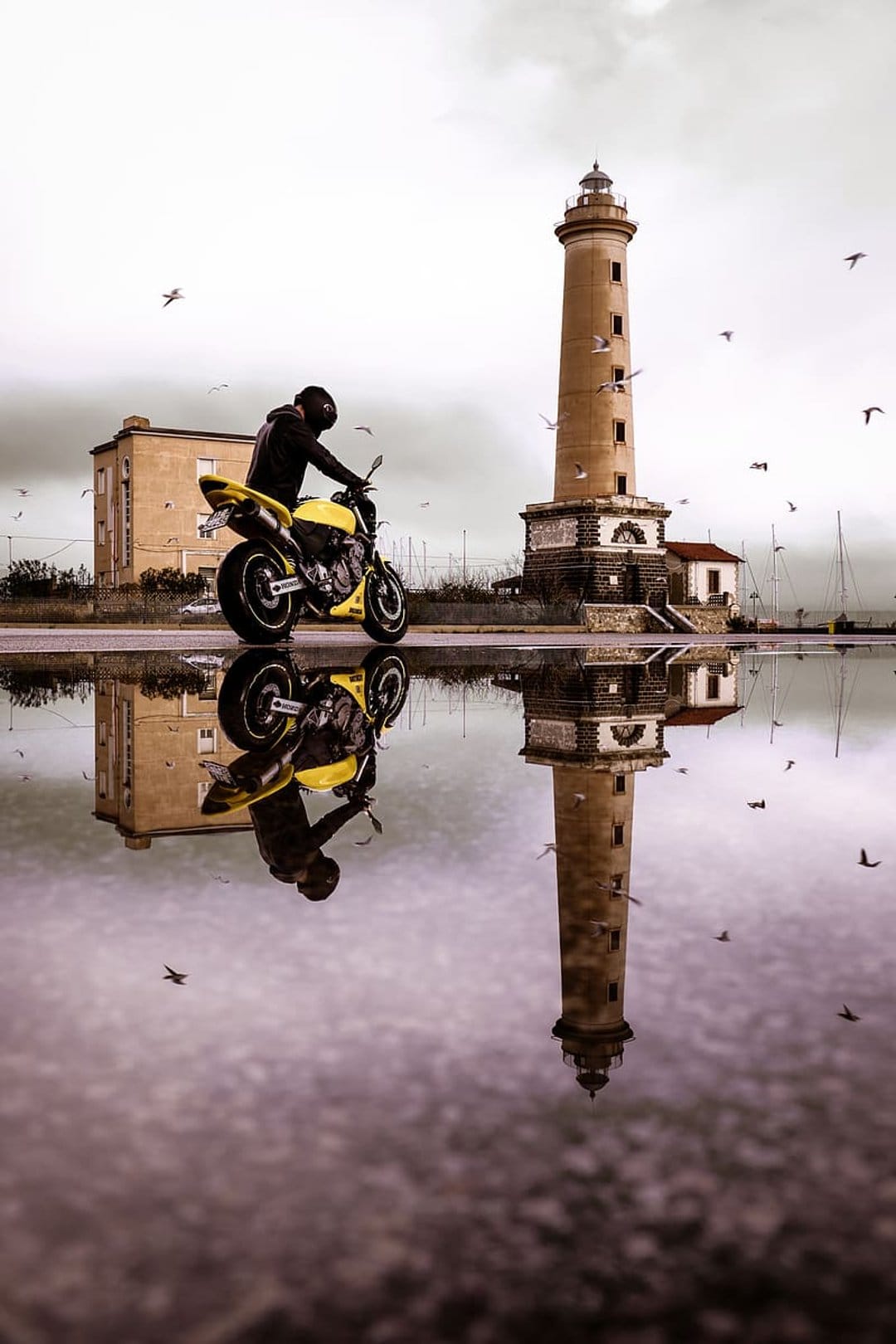 Motorcyclist with a yellow bike reflected in water beside a tall lighthouse under a bird-filled overcast sky.
